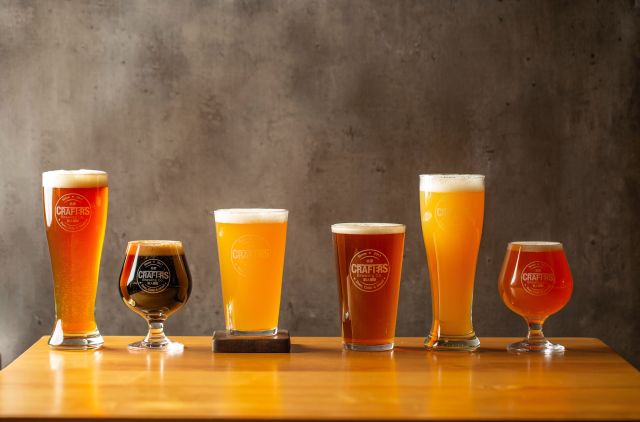 six glasses of varying sizes full of craft beer in a range of colors of red, gold and brown.
