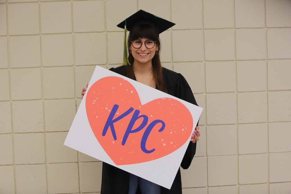 Woman with brown hair and glasses in graduation cap and gown holding KPC sign