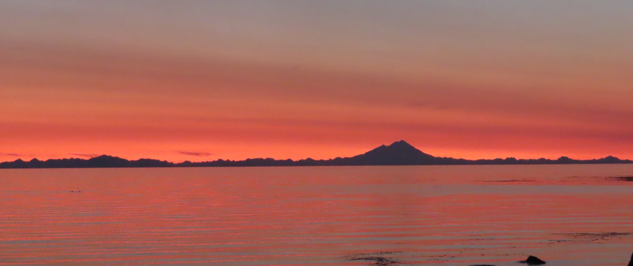 a volcano on the horizon of a body of water with a colorful sunset.