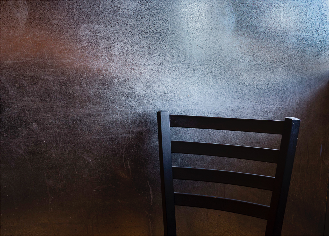 The back of a wooden chair set against a cement looking wall a faint shaft of sunlight falls over the top of the chair and is the only light in the room 