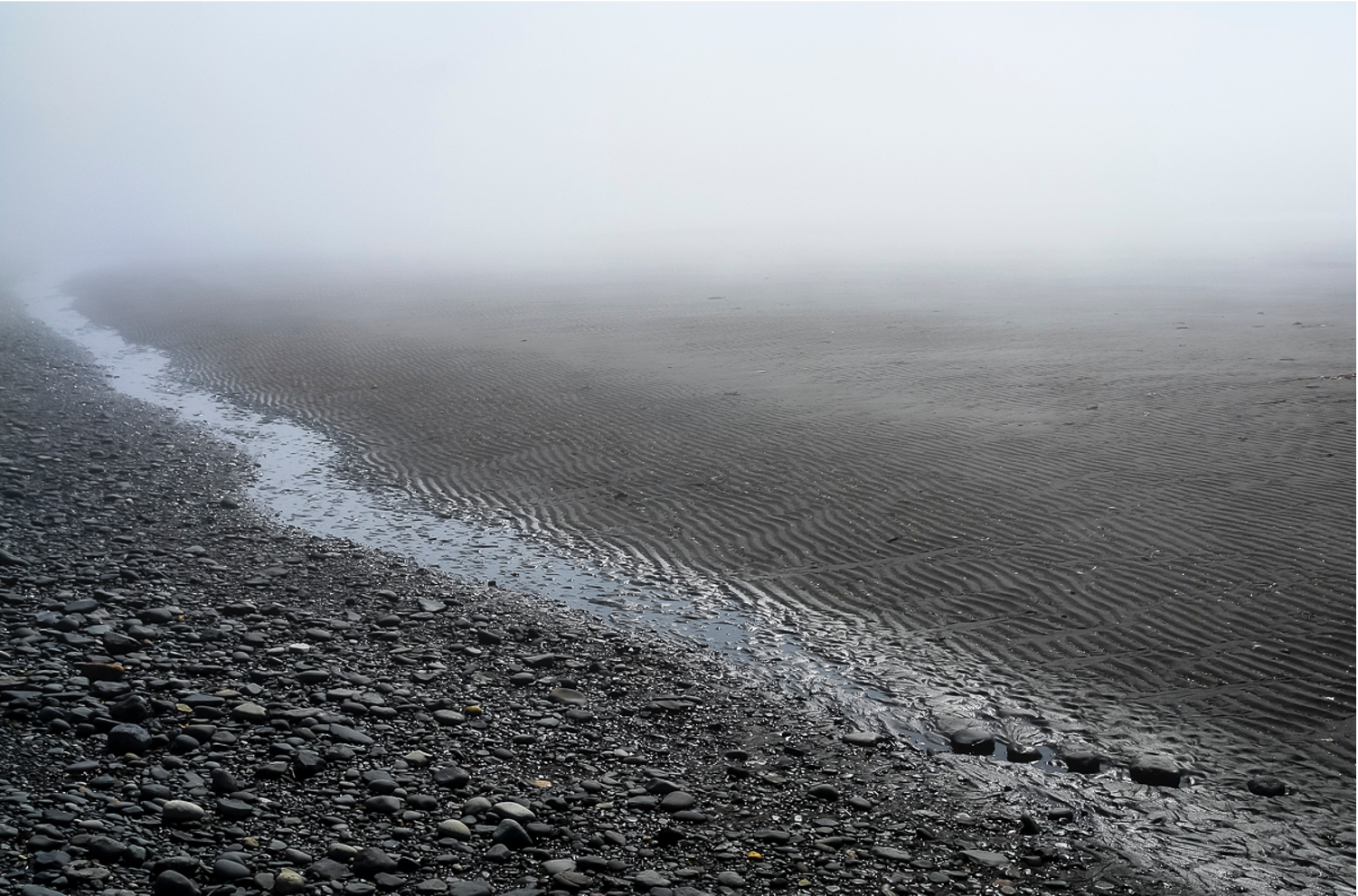 a rocky beach beach that leads into a sandy beach the day has fog and is gray. A line of water seems to seperate the rocky and sandy areas