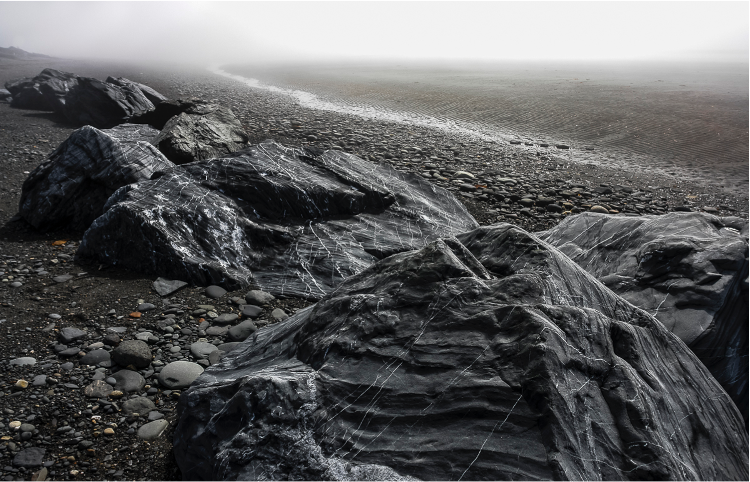 Large rocks in the sand and pebbles at a beach on a gray day with fog